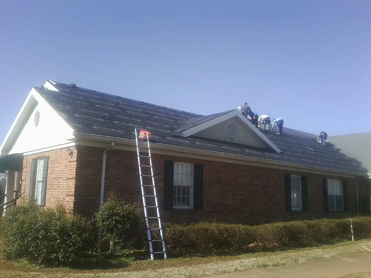 Crew tearing shingles off a brick ranch roof with a ladder set — Metal Roof Repair work by our crew in Kittrell
