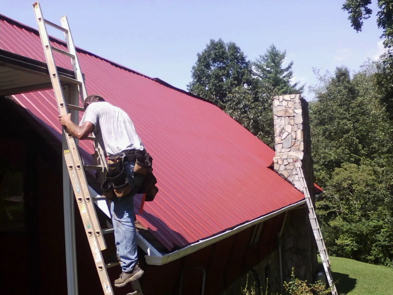 Roofer on a ladder beside a red metal roof and stone chimney — Soffit & Fascia Repair by Roofing Brothers in Kittrell